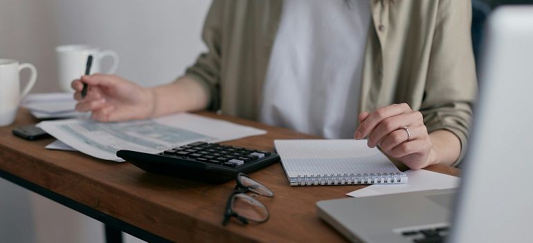 Person working at a desk with a calculator, notebook, and laptop in a bright office