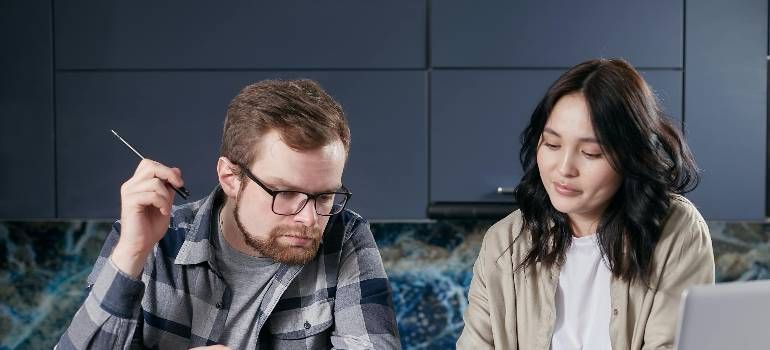 Two coworkers sit at a desk, looking at a laptop and discussing something with focused expressions.