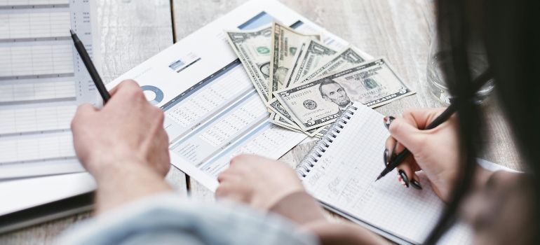 Hands writing on financial forms beside stacked dollar bills on a desk