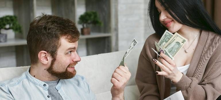 Man handing cash to woman while she smiles and holds a fan of money indoors