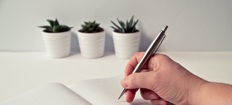 Hand writing in an open notebook beside three small potted plants on a white desk
