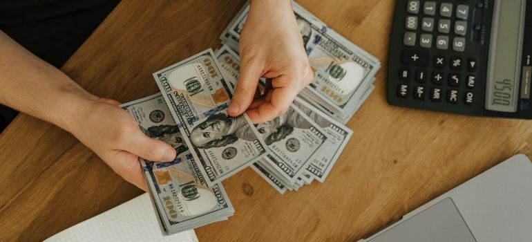 Hands counting U.S. dollar bills on a wooden desk beside a calculator and laptop keyboard