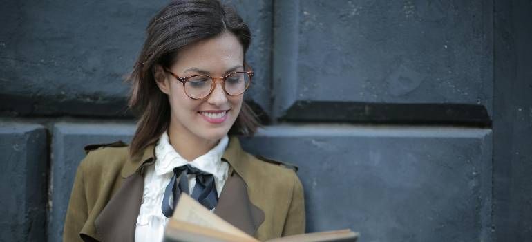 Woman in glasses reading a book against a dark wall, wearing a tan blazer and white blouse.