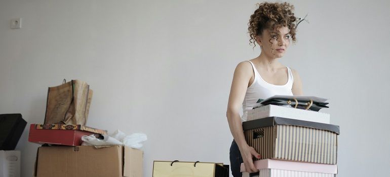 Woman carrying a stack of boxes in a sparsely furnished room, with more boxes and bags nearby