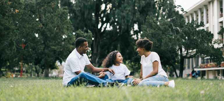 Three people sitting on grass in a park, talking and smiling near trees and a building.