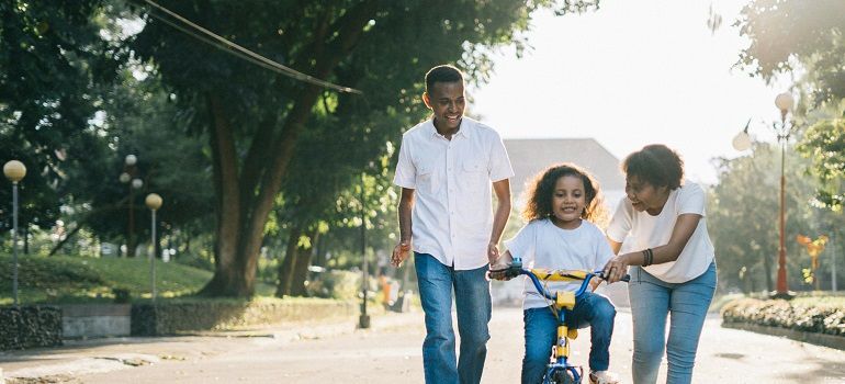 Family helping a child ride a bicycle on a tree-lined path at sunrise