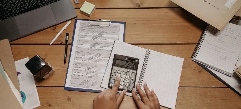 Hands using a calculator on paperwork at a wooden desk with notebooks and office supplies