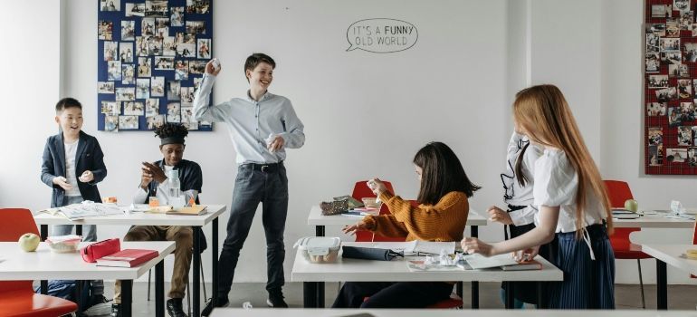 Office meeting with people discussing around tables, one person presenting by a whiteboard.