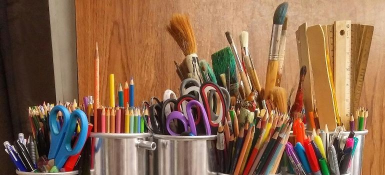 Assorted paintbrushes, pencils, scissors, and art tools in containers against a wooden background