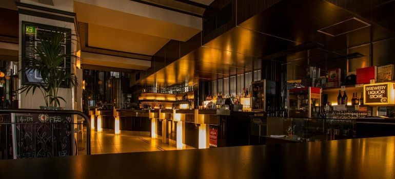 Dimly lit modern bar interior with wooden tables, shelves of bottles, and warm amber lighting
