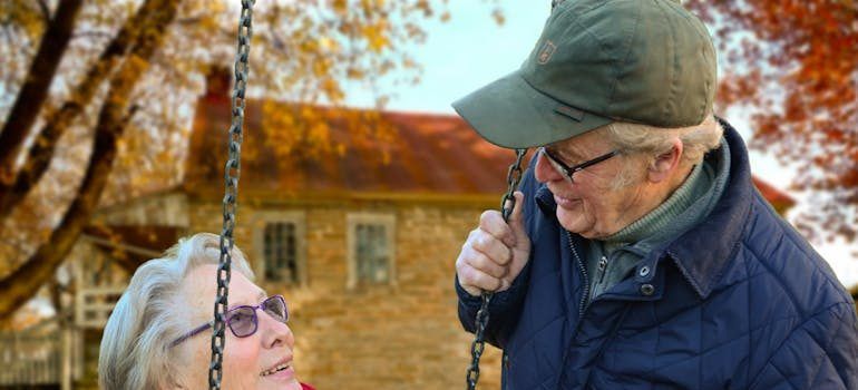 Man in cap and glasses gently pulling a child on a swing outdoors near a brick building, autumn colors visible