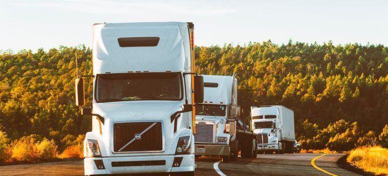 Three white semi-trucks driving on a curving highway beside autumn trees