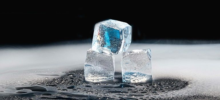 Three ice cubes on a wet black surface, one glowing blue in dramatic light.