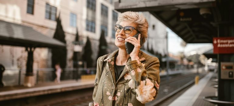 Woman in glasses talks on a phone at a train platform, with a blurred station in the background