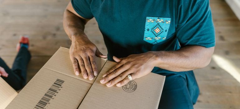 Person in teal shirt taping closed a cardboard box on a wooden floor