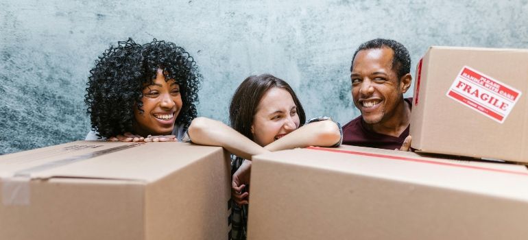 Three smiling people peeking over moving boxes, one labeled “FRAGILE”