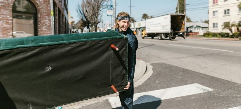 Person carrying a large green tarp across a street near parked trucks and apartment buildings