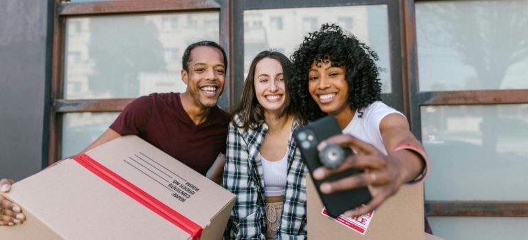 Three smiling people holding moving boxes and a camera on a porch step outdoors