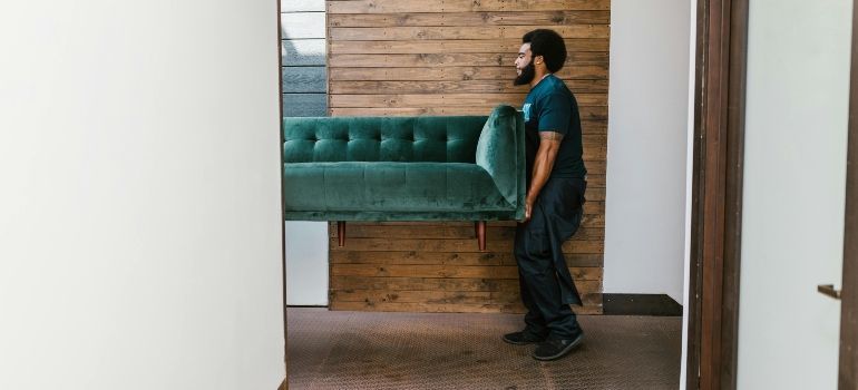 Person carrying a green sofa through a doorway in a hallway with wooden walls and floor