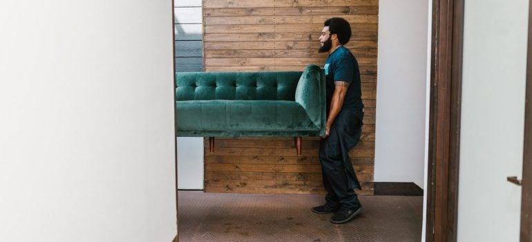 Person carrying a teal sofa through a doorway in a modern hallway