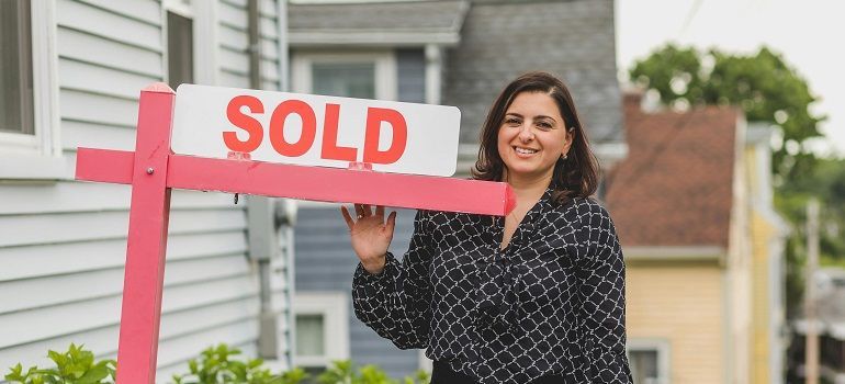 Smiling woman holding a pink SOLD real estate sign outside a house