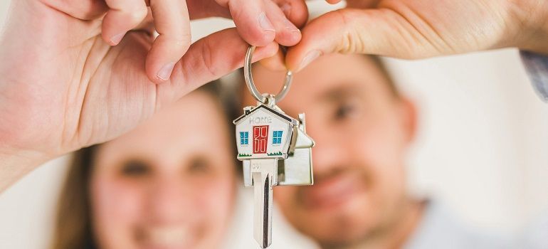 Hands holding a house keychain with a blurred couple in the background