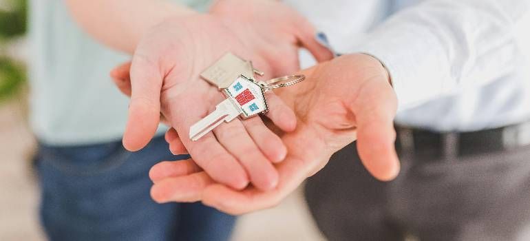 Hands holding a small house keychain with a tiny red-and-white house charm over a blurred background
