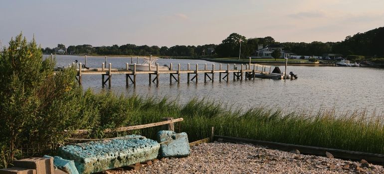 Wooden dock extending into a calm lake beside a pebble shore and grassy bank, with teal benches in the foreground