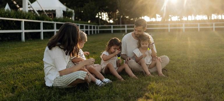 Family sitting on grass at sunset, feeding a small child outdoors near a white fence.