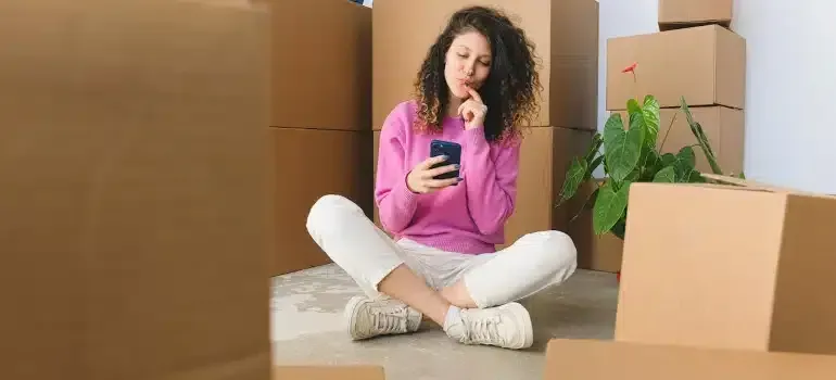 Woman sitting among moving boxes, looking at her phone and smiling in a room with a potted plant.