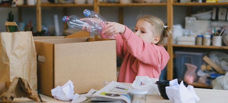 Child reaching into a cardboard box amid craft supplies on a cluttered table