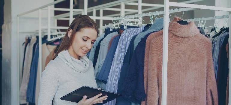 Woman browsing clothes on a rack in a shop, holding a tablet and examining a pink sweater.