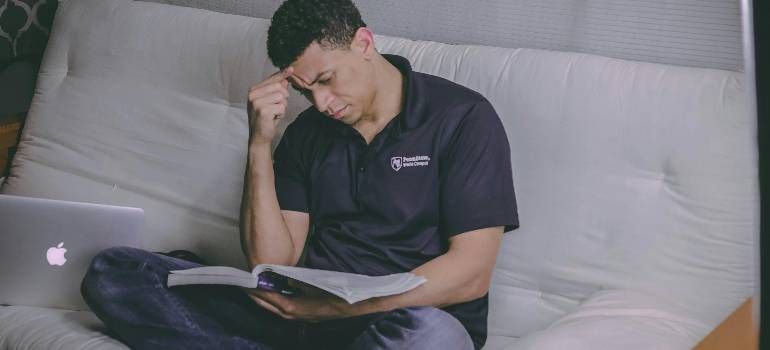 Person sitting on a couch, reading a book beside an open laptop with a thoughtful expression.