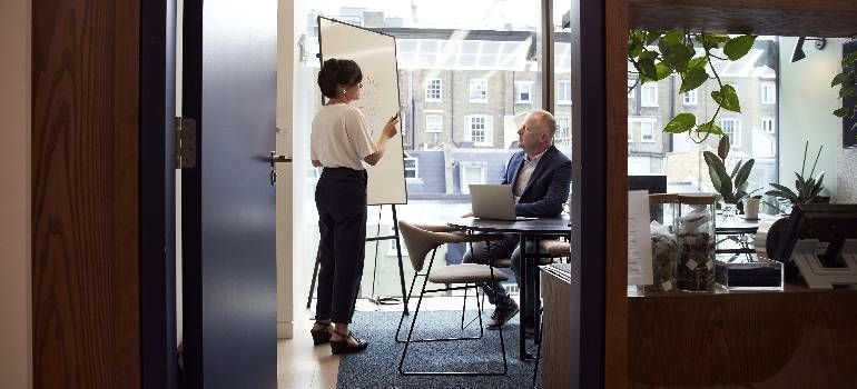 Two people talking in a bright office near a laptop and large windows.