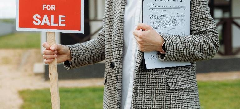 Real estate sign saying “For Sale” held beside a person holding a house listing