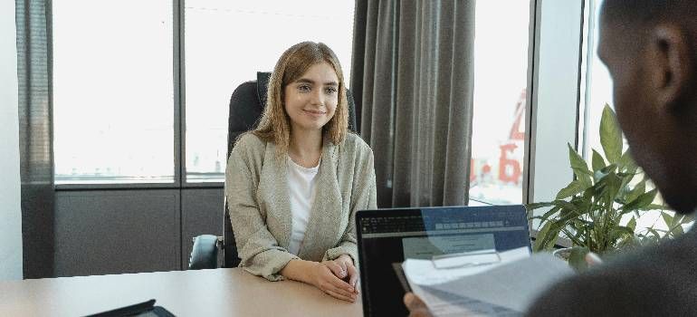 Two people in an office meeting, with a woman smiling across a laptop and papers in view