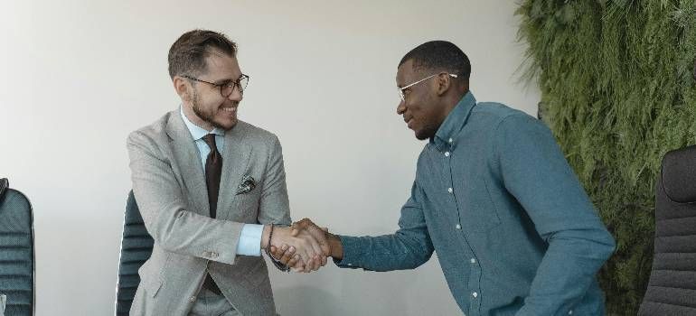 Two men shake hands in an office, one in a gray suit and one in a blue shirt.