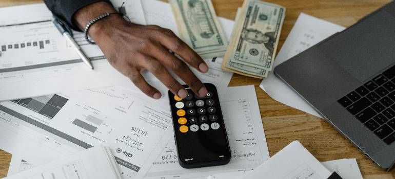 Hand using calculator on bills and paperwork beside laptop and cash on a desk