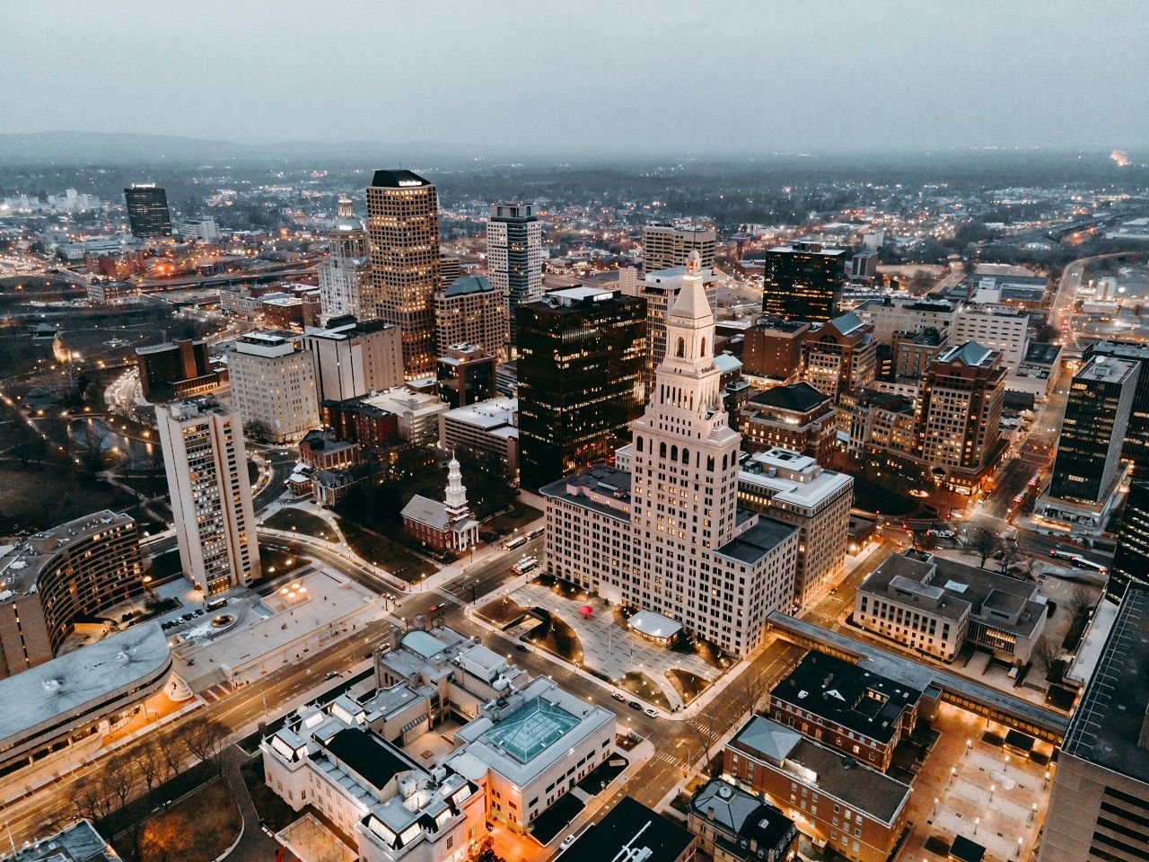Aerial view of a city skyline at dusk with lit buildings and a prominent white tower in the center