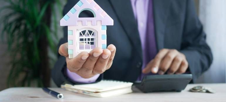 Hand holding a pink house model next to a calculator and notebook on a desk