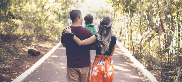 Two adults and a child walking on a sunlit path through trees, seen from behind.
