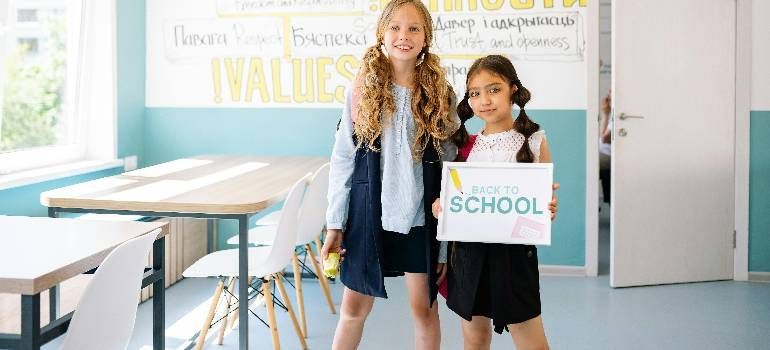 Two children stand in a bright classroom holding a “Back to School” sign.