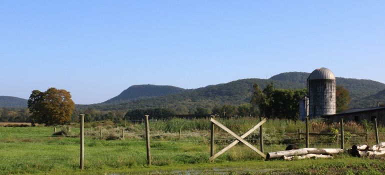 Rural pasture with fence posts, a gate, green fields, wooded hills, and a silo under a clear sky