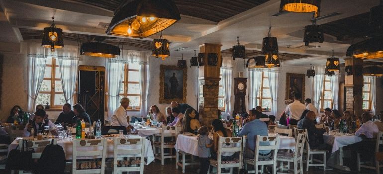 Crowded restaurant dining room with people seated at white tables under warm pendant lights