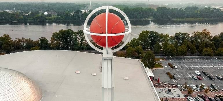 Large red spherical sculpture atop a building, overlooking trees, river, and parking lot