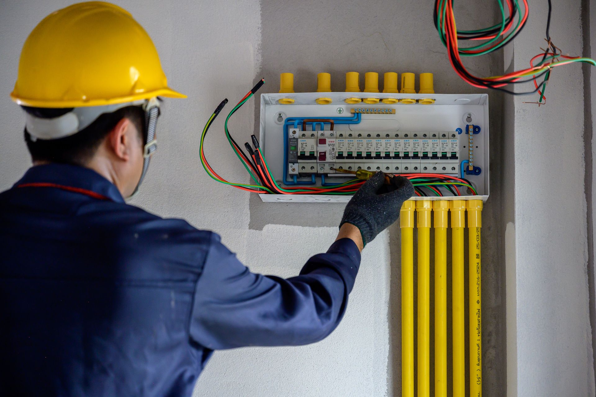 Electrician in a blue jumpsuit and hard hat working on a circuit breaker box on a gray wall.