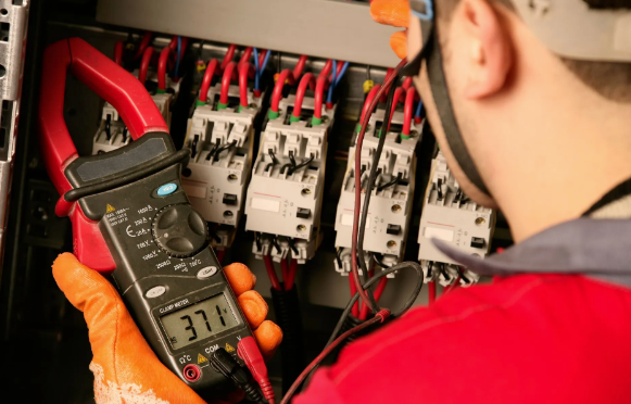 Electrician using a clamp meter to measure current in an electrical panel, wearing a hard hat and gloves.