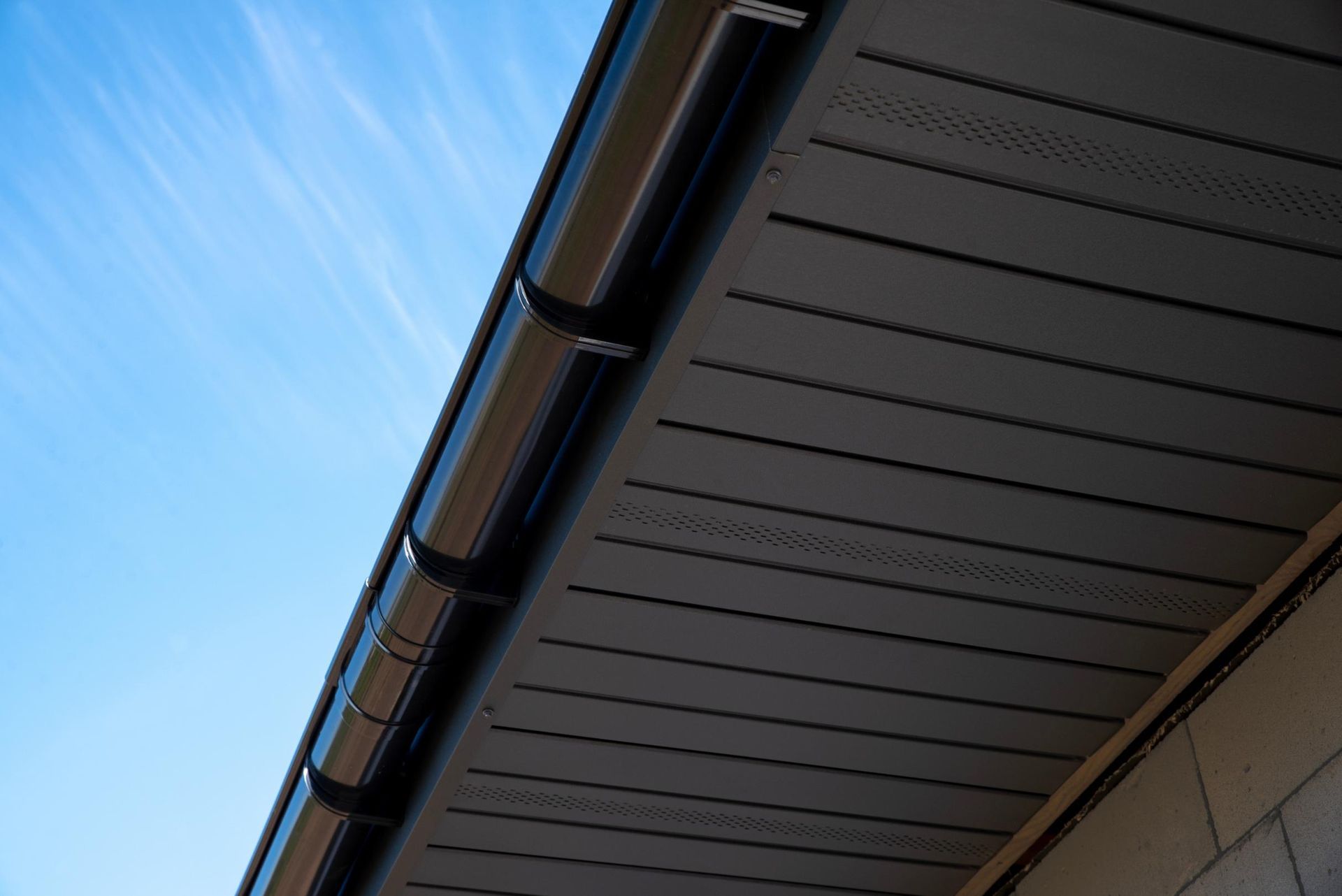 A low-angle view of a house roof eaves, featuring dark brown siding and a black gutter against a clear blue sky.