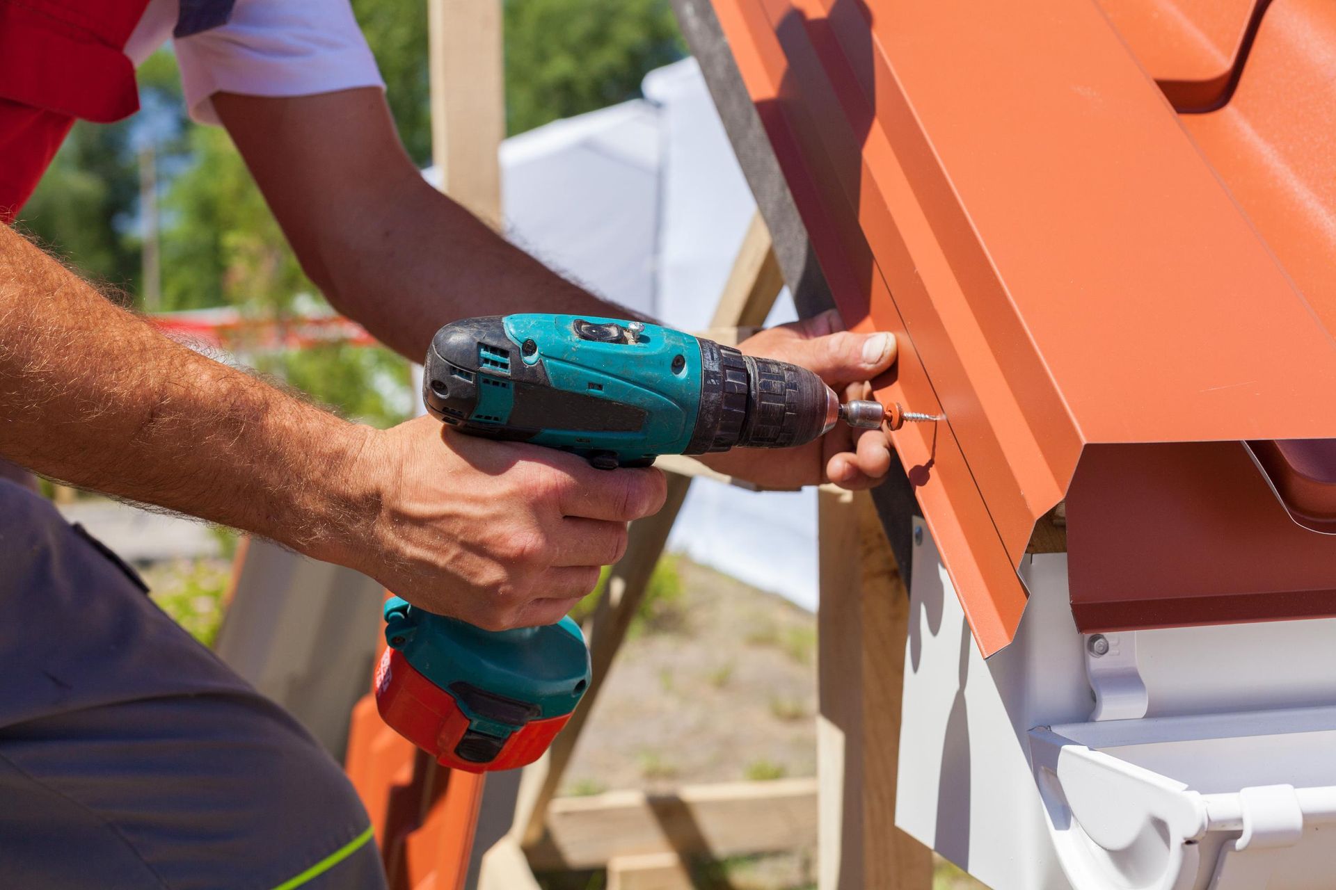 A construction worker using a cordless drill to secure orange metal roofing panels onto a wooden structure.