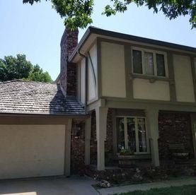 A two-story Tudor-style house featuring a brick chimney, a wood-shake roof, beige stucco siding, and a white garage door.